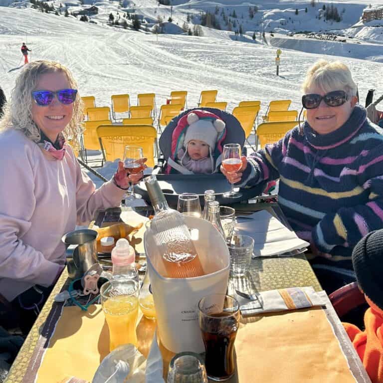 Three generations—woman, baby, and older woman—cheers with glasses of rosé wine at an outdoor ski resort table in La Plagne, surrounded by snow and yellow chairs.