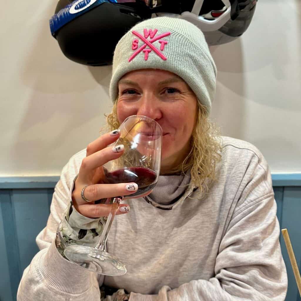 Smiling woman in a winter hat with a pink ski logo sips red wine in a cozy après-ski setting, with ski gear hanging on the wall behind her.