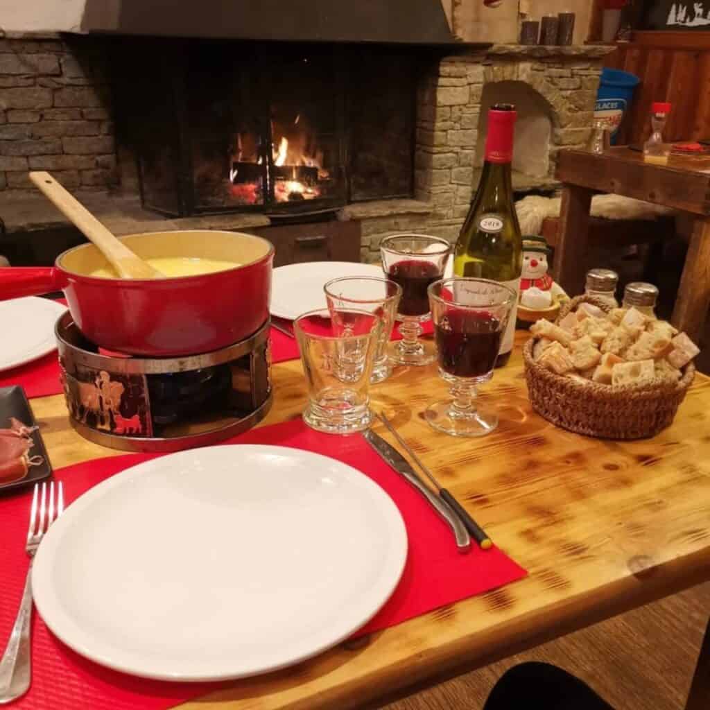 Rustic alpine dinner setup with a pot of cheese fondue, red wine in goblets, and a bottle of Savoie wine near a crackling fireplace.