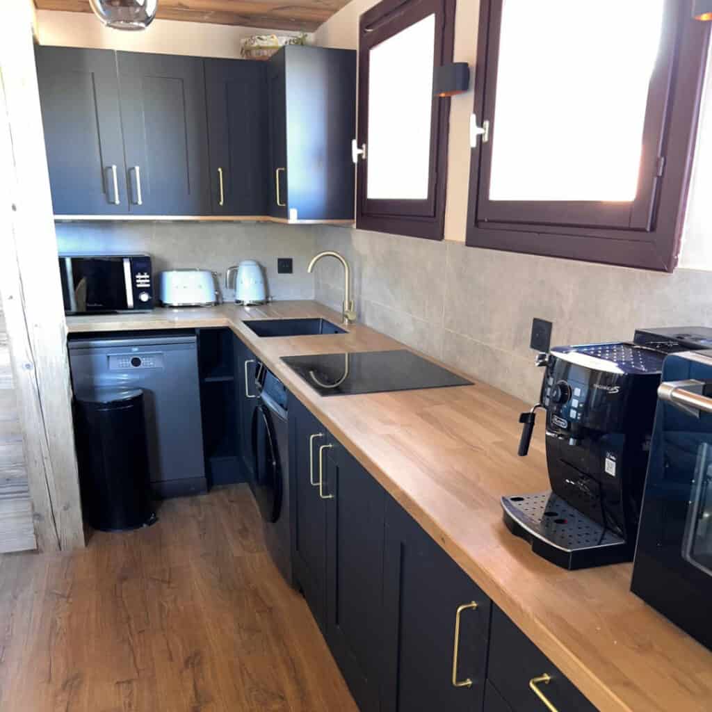 Modern chalet kitchen with matte black cabinets, wood countertops, and brass accents, featuring a coffee machine, toaster, induction hob, and built-in appliances under large sunlit windows.