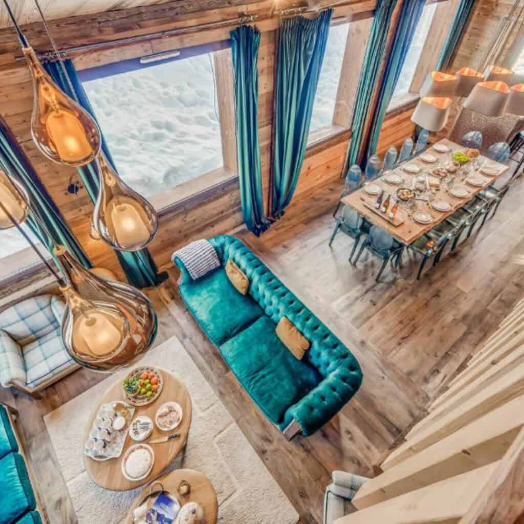 Overhead view of a luxury chalet’s living and dining space, featuring emerald green sofas, glowing pendant lights, and a long wooden dining table set for guests by snow-lined windows.