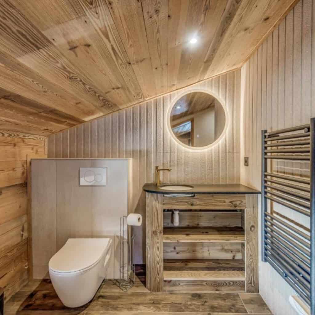 Stylish chalet bathroom with wood-panelled sloped ceiling, a modern wall-mounted toilet, a black countertop vanity with brass tap, and a round backlit mirror.