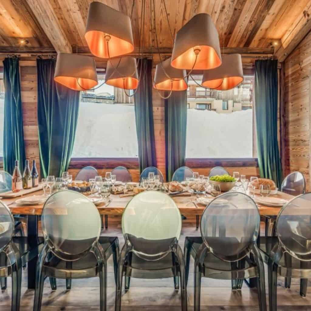 Close-up of a long chalet dining table set for a meal with wine, water glasses, and fresh bread, surrounded by clear chairs and framed by rich green curtains and wood-clad walls.