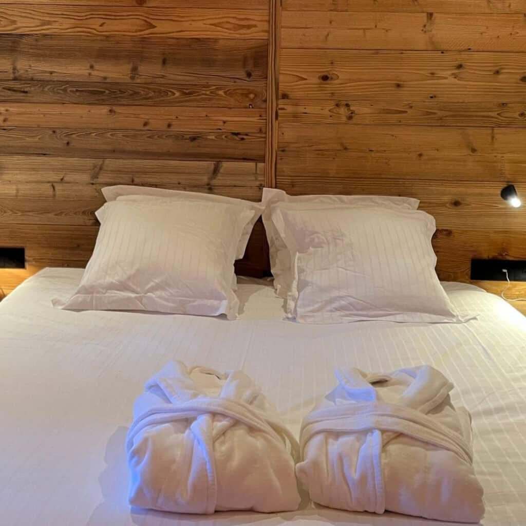 Cozy chalet bedroom with wood-paneled walls, crisp white bedding, and two fluffy bathrobes neatly placed on the bed under warm lighting.