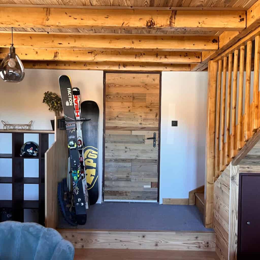 Rustic chalet entryway with wood detailing, stairs leading up, and ski gear including skis, a snowboard, and a helmet stored neatly beside the front door.