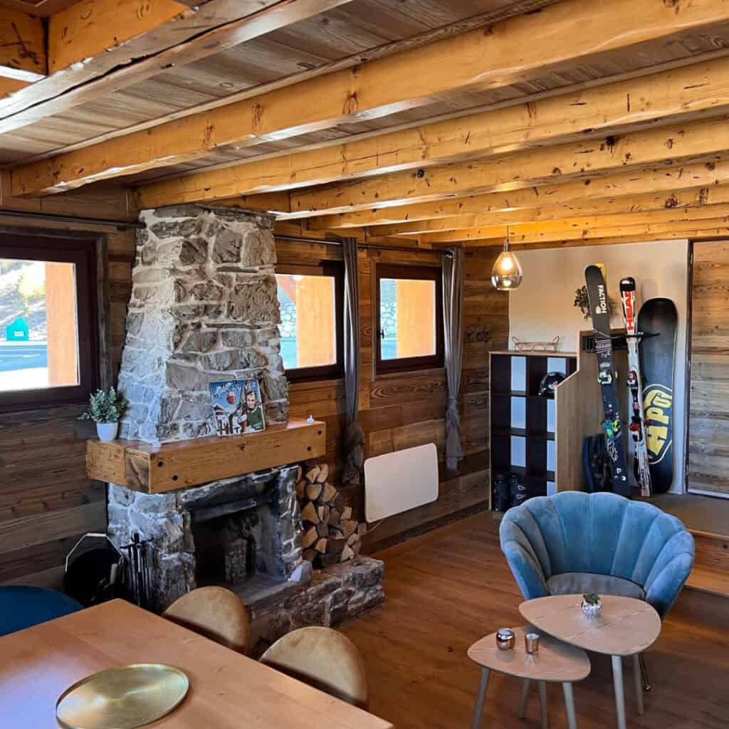 Inviting chalet living room with exposed wooden beams, a stone fireplace stocked with logs, ski equipment storage, and stylish modern furnishings including a velvet chair and nested coffee tables.