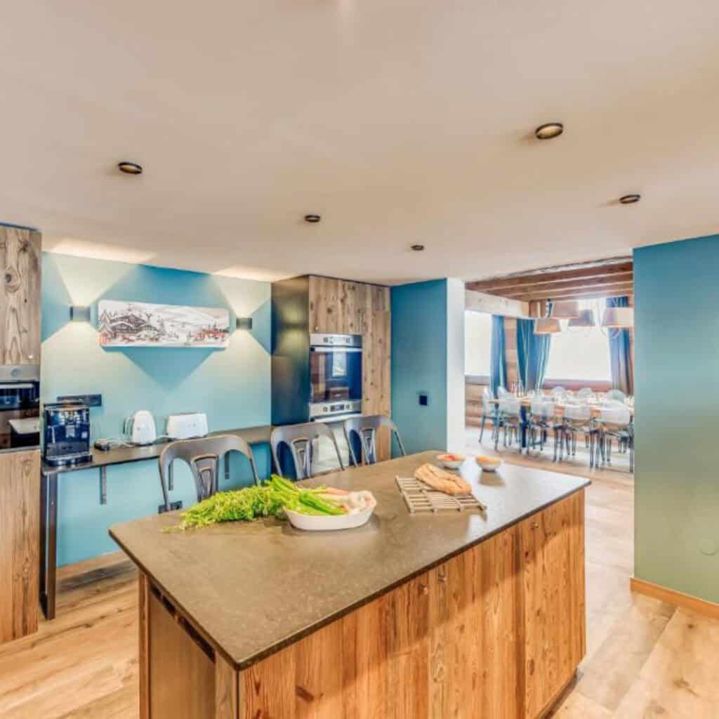 Open-plan chalet kitchen with a rustic wood island, sleek modern appliances, and a view into a stylish dining area with blue walls and warm lighting.