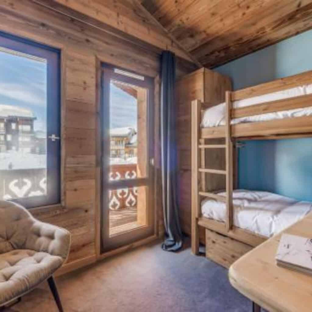 Bright chalet bedroom featuring wooden bunk beds, a reading chair, and views of snow-covered chalets through large windows and a private balcony door.