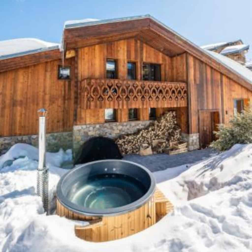 Chalet exterior with natural wood cladding and a balcony, shown on a sunny winter day with a steaming round outdoor hot tub nestled in the snow.