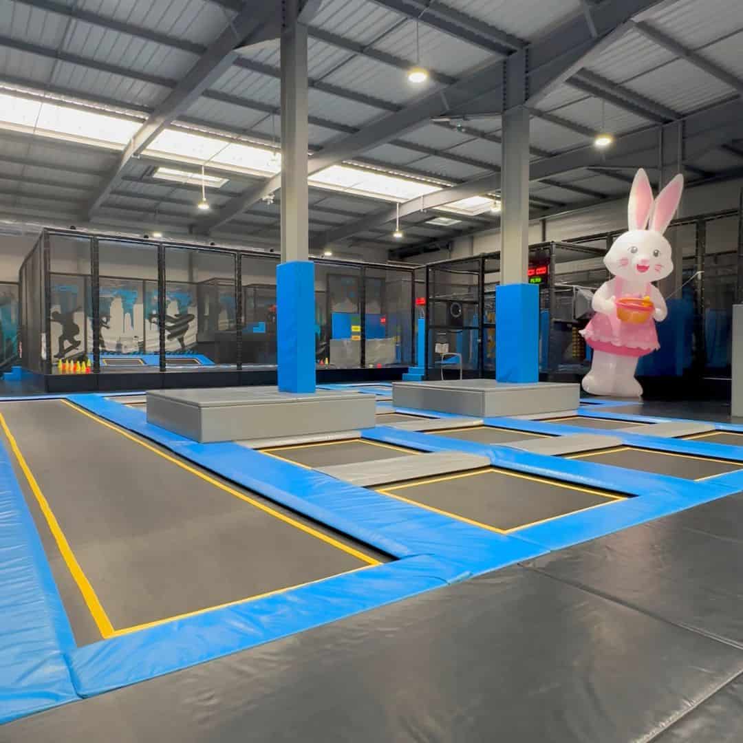 Indoor trampoline park in Albertville with rows of blue and black trampolines and padded platforms. A large bunny mascot stands in the corner showing a fun stop on a family day trip La Plagne.