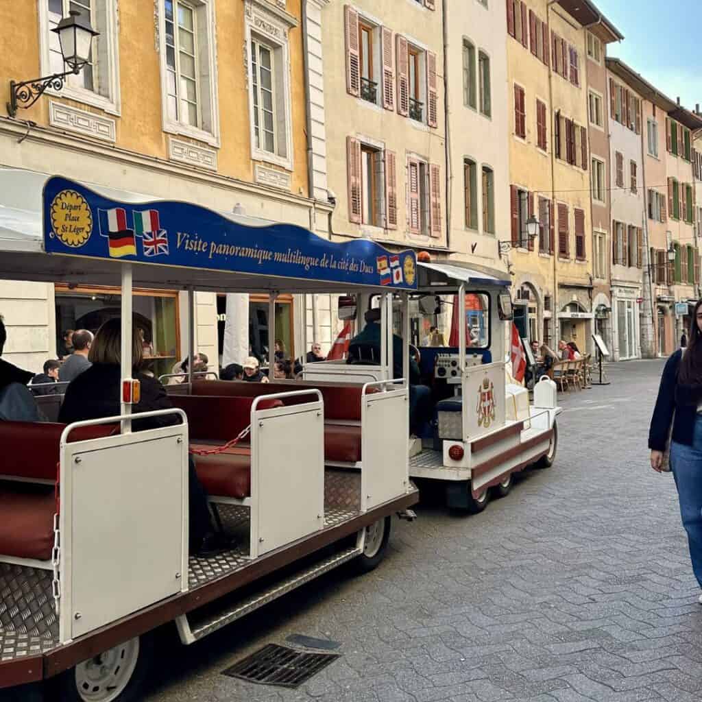 Small tourist train driving through a charming street in Chambéry lined with pastel buildings and shuttered windows. A relaxed way to explore the old town on a family day trip from La Plagne.