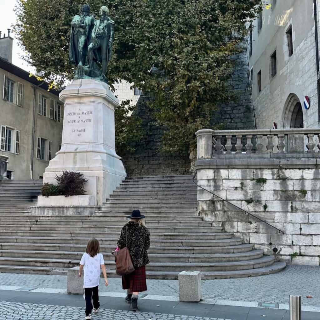 Adult and child walking toward a large statue at the top of stone steps in Chambéry old town. Exploring historic streets on a family day trip French Alps.