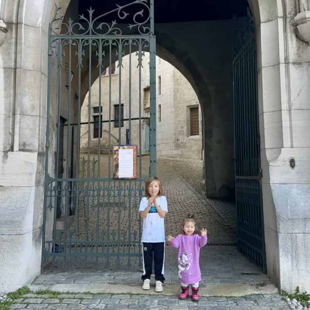 Two children standing in front of a gated stone archway leading into a quiet courtyard. Discovering hidden corners during a day trip from La Plagne.