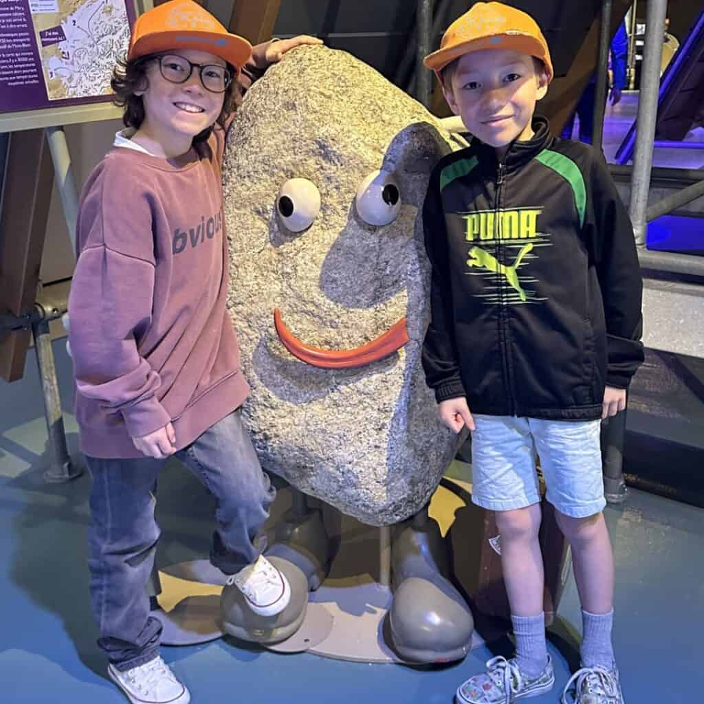 Two kids posing beside a playful rock sculpture with a smiling face inside a museum. Fun interactive exhibit during a family day trip French Alps.