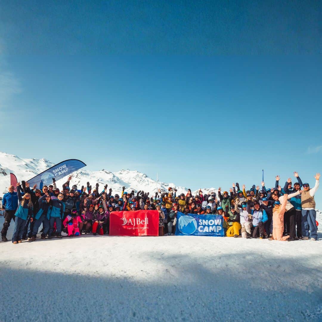 Large group of skiers and snowboarders stand on a snowy mountain holding Snow Camp and AJ Bell banners with arms raised. This wide group photo captures the energy of the Snow Camp 3 Valley Rally in a bright alpine setting.
