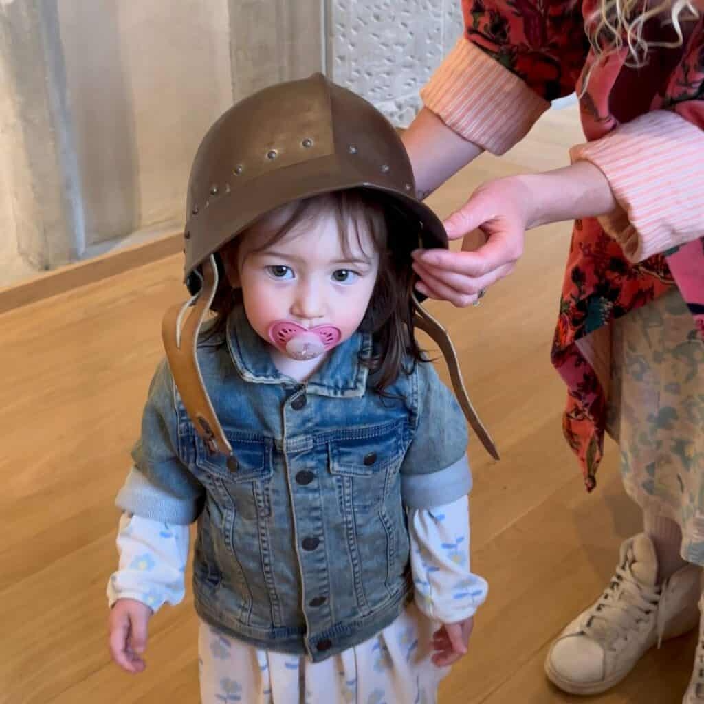 Young child wearing a medieval helmet being adjusted by an adult inside a museum. Playful history experience during a family day trip French Alps.