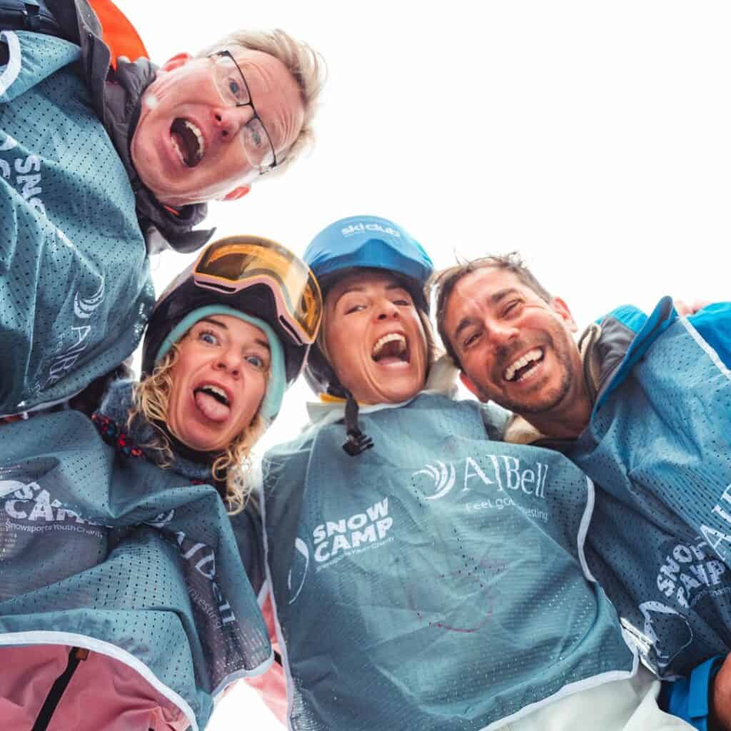 Four adults in Snow Camp bibs lean in close smiling and pulling playful faces in ski gear. This candid moment shows the fun and team spirit at the Snow Camp 3 Valley Rally.
