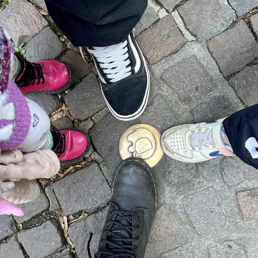 Four pairs of feet gathered around a brass sidewalk marker set into cobblestones in Chambéry. Close up moment highlighting a fun detail to spot during a family day trip French Alps.