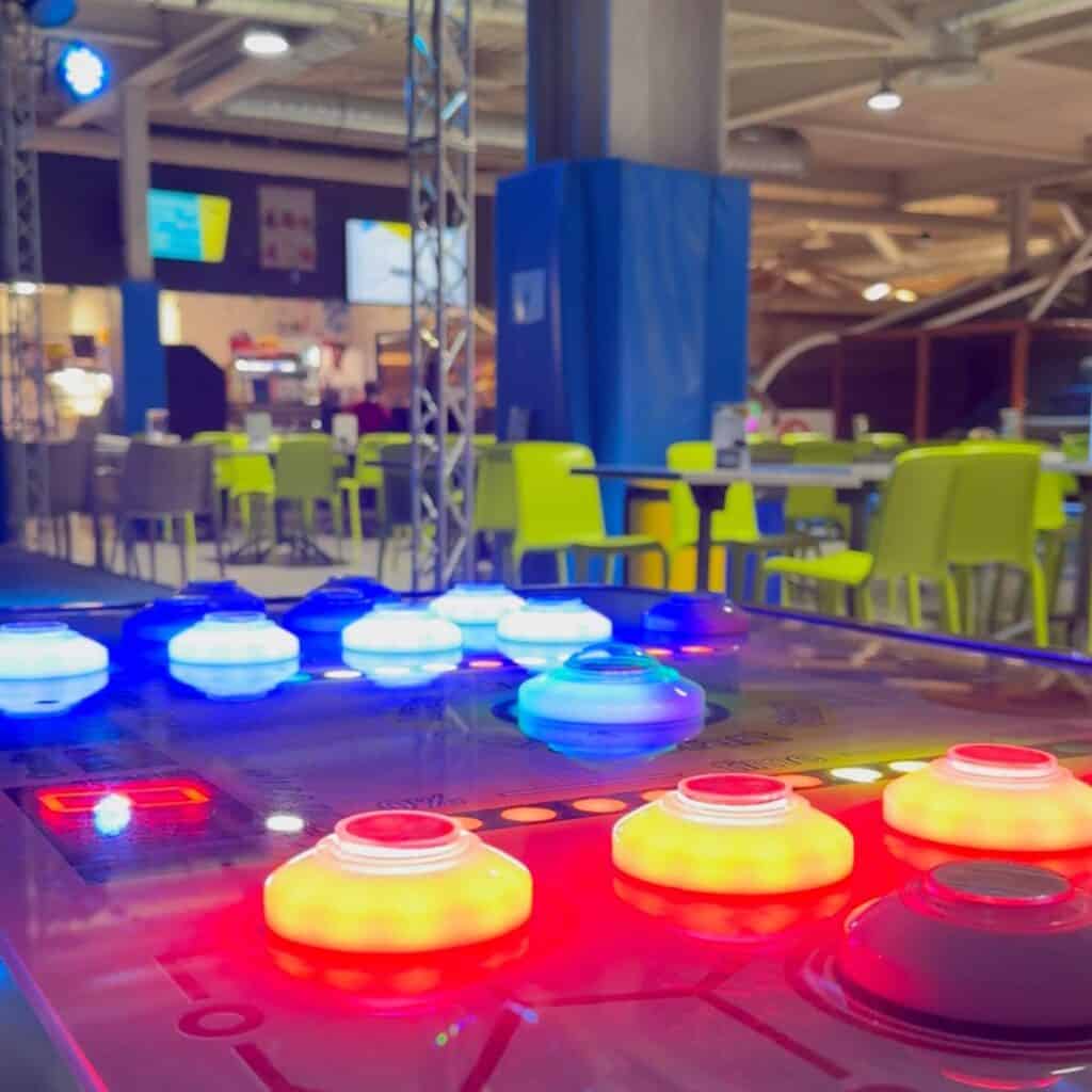 Arcade game table glowing with red blue and yellow lights in a seating area with bright green chairs. Entertainment zone at a trampoline park Albertville offering a break between activities.