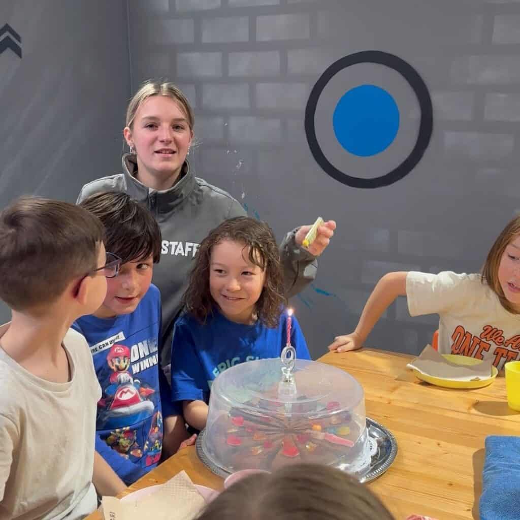 Kids gathered around a birthday cake with a lit candle while a staff member stands behind them. Celebration moment at K Park Albertville during a family day trip La Plagne.