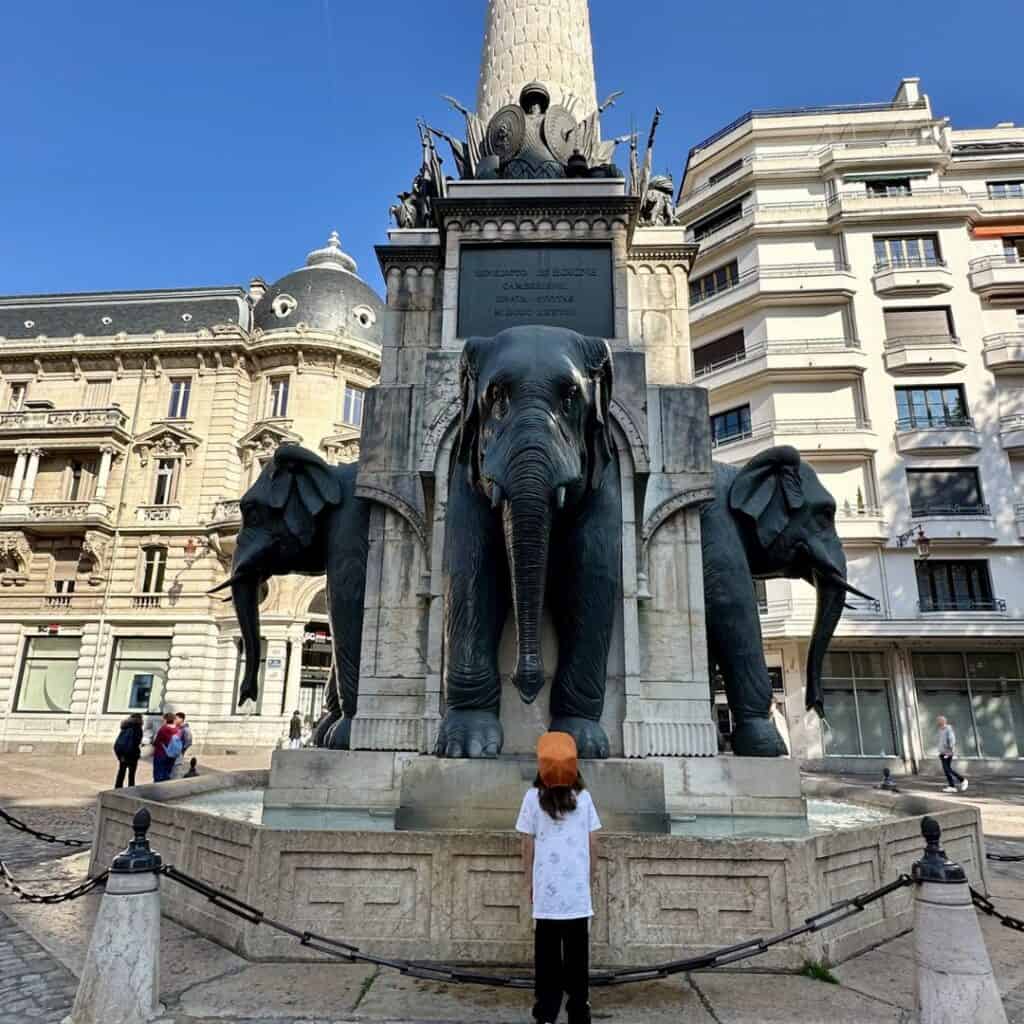 Child looking up at the famous elephant fountain in Chambéry surrounded by historic buildings. Iconic landmark to visit during a family day trip French Alps.