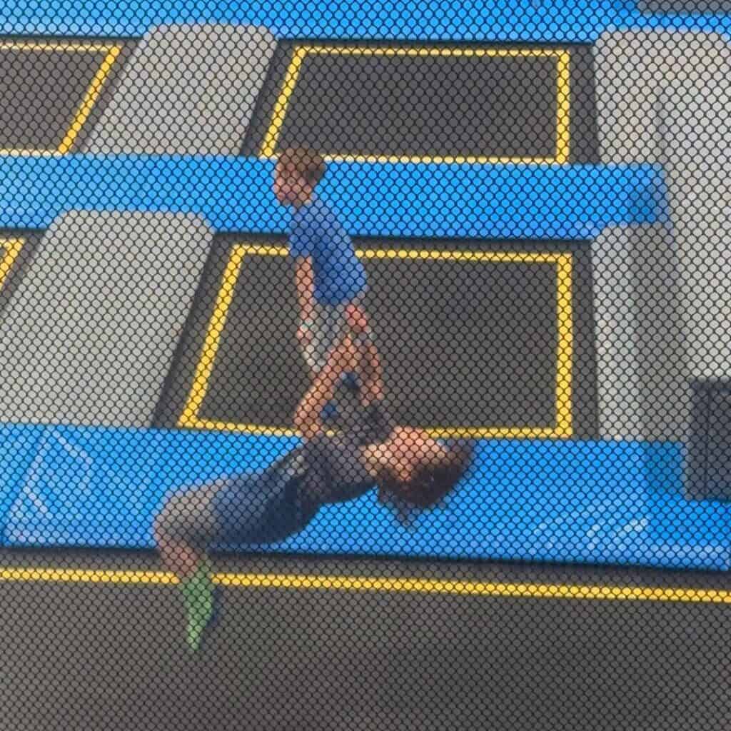 Child mid flip on a trampoline while another stands nearby on padded blue and black jumping lanes. Action shot showing the excitement of a trampoline park Albertville.