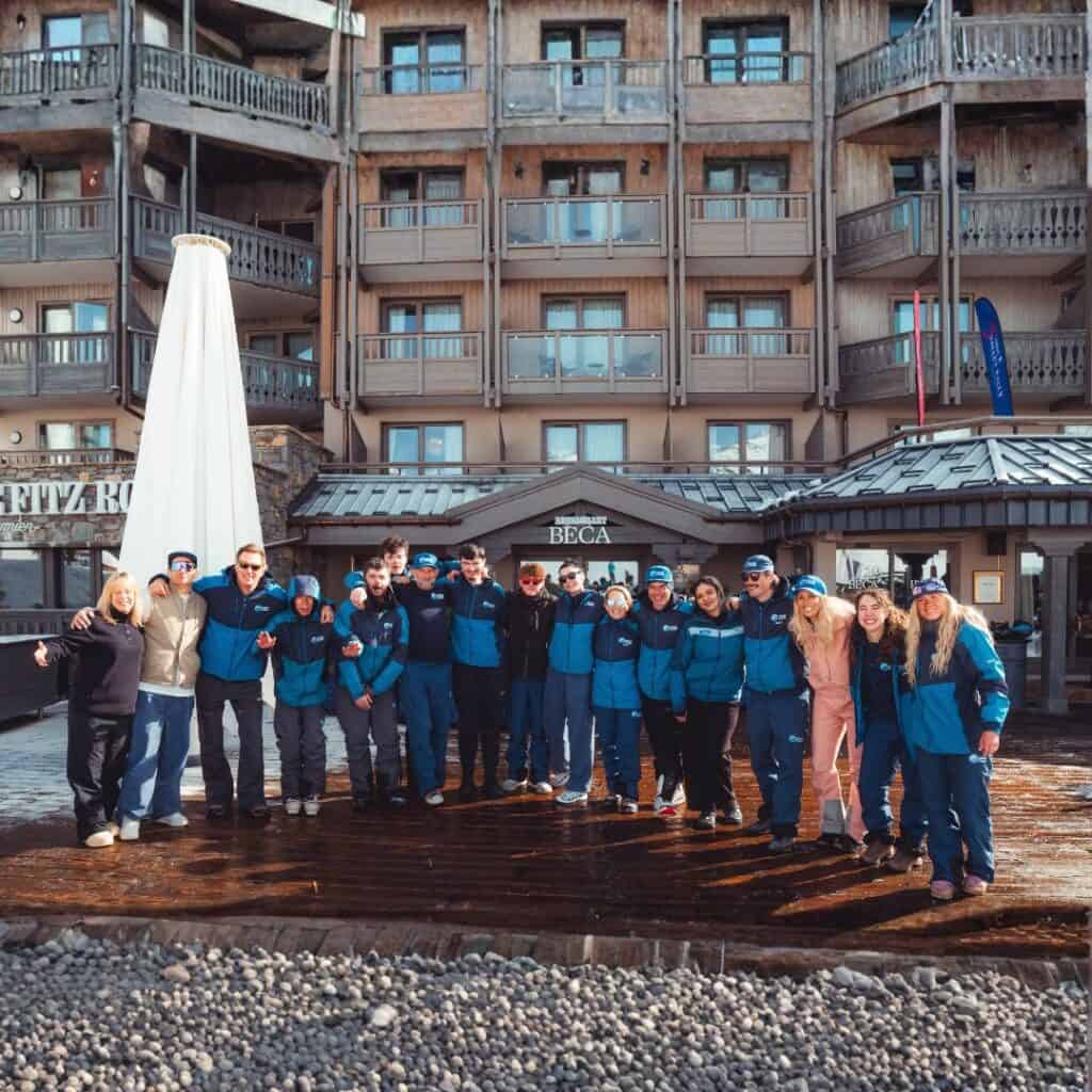 Group of Snow Camp staff and participants stand together outside a chalet style hotel smiling arm in arm. This team photo captures connection and community at the Snow Camp 3 Valley Rally.