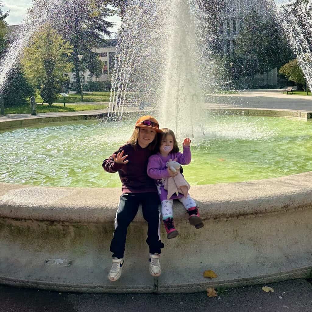 Two children sitting on the edge of a fountain smiling with water spraying behind them in a sunny park. Relaxed moment during a family day trip French Alps.