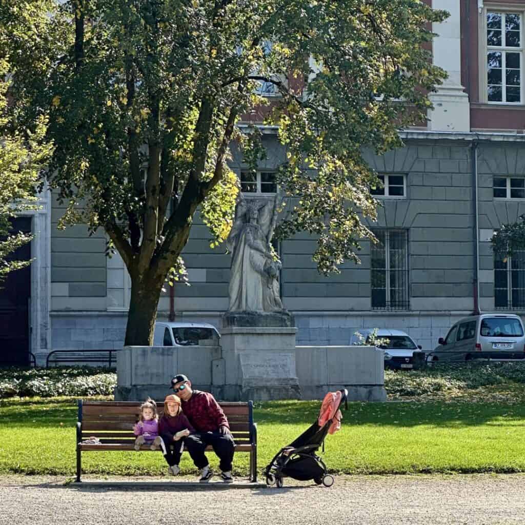 Family sitting on a park bench with a stroller nearby under leafy trees in Chambéry. Peaceful break during a family day trip French Alps.