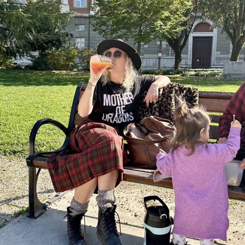 Woman sitting on a bench drinking from a bottle while a child stands beside her in a sunny park. Casual moment enjoying a day trip from La Plagne.