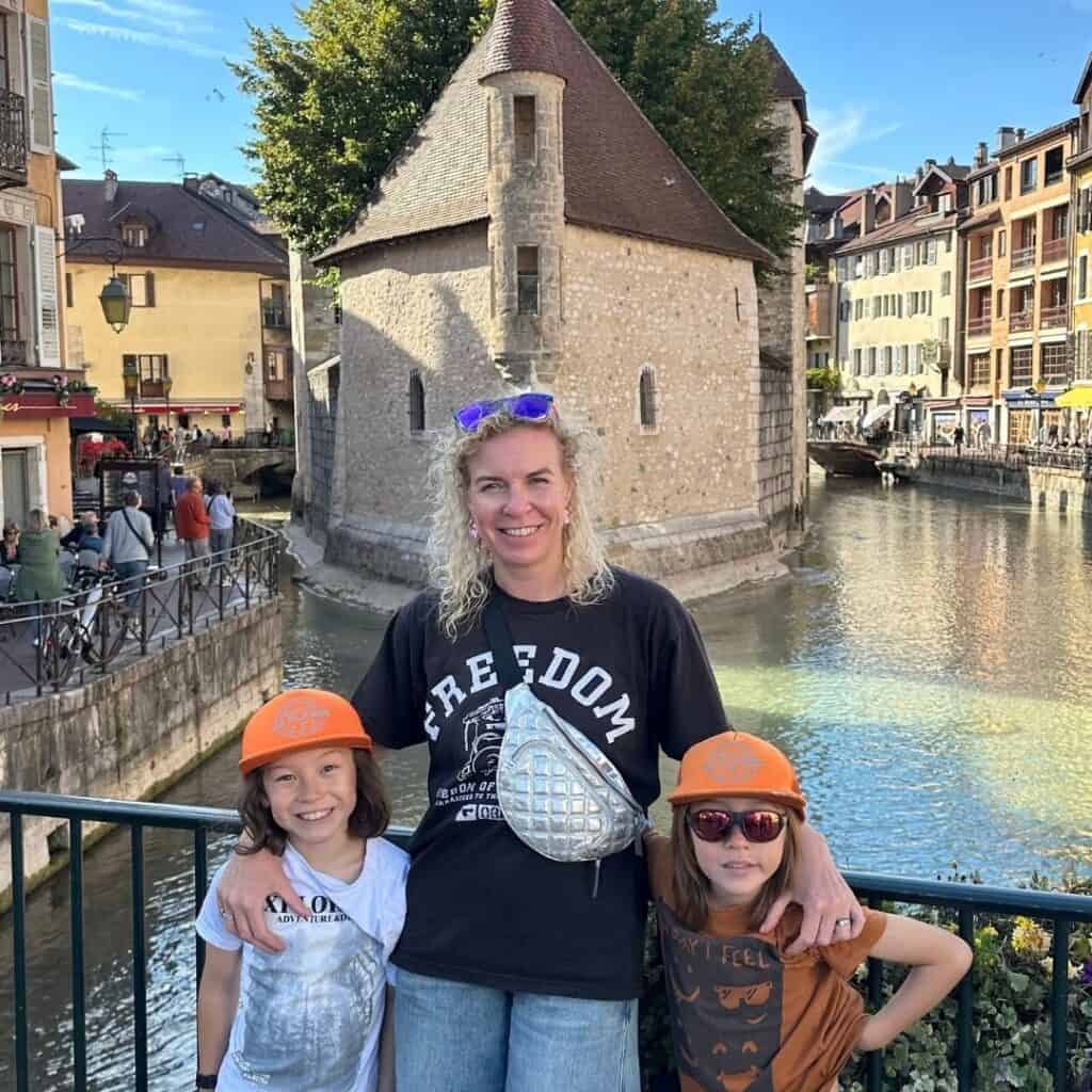 Woman standing with two children in front of the historic Palais de l’Isle building in Annecy surrounded by canals and pastel-colored buildings. The family poses on a sunny day while exploring one of the most popular day trips from La Plagne.