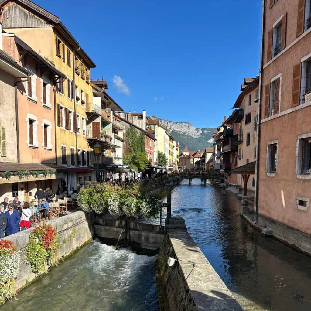 Colorful buildings line the canal in Annecy with outdoor cafés, flower boxes, and mountain peaks in the distance under a bright blue sky. The scenic old town is a popular day trip from La Plagne and a classic stop in the French Alps and Savoie region.