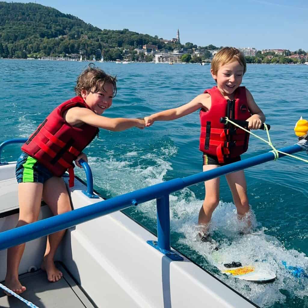 Two children wearing red life jackets hold hands while wakeboarding beside a boat on the turquoise water of Lake Annecy. Forested hills and lakeside villages in the French Alps rise in the background on a sunny summer day.