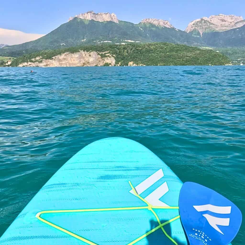 View from a blue paddleboard floating on Lake Annecy with mountain cliffs and green hills surrounding the water in the French Alps. A blue paddle rests on the board beneath a clear summer sky in the Savoie region.