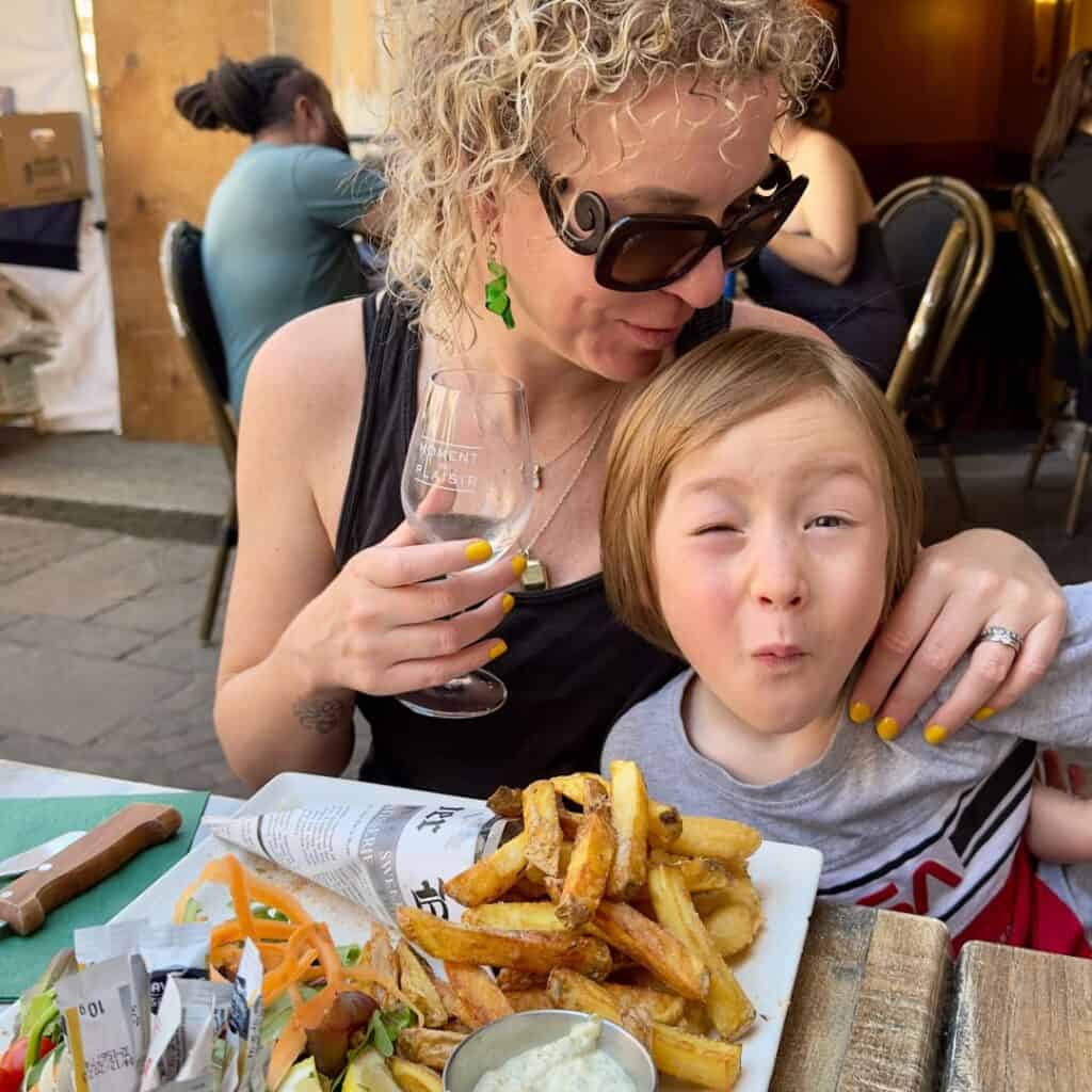 Woman with curly blonde hair and sunglasses sits beside a child making a funny face at an outdoor restaurant in Annecy. A plate piled with thick-cut fries and salad sits on the table during a relaxed lunch in the French Alps.