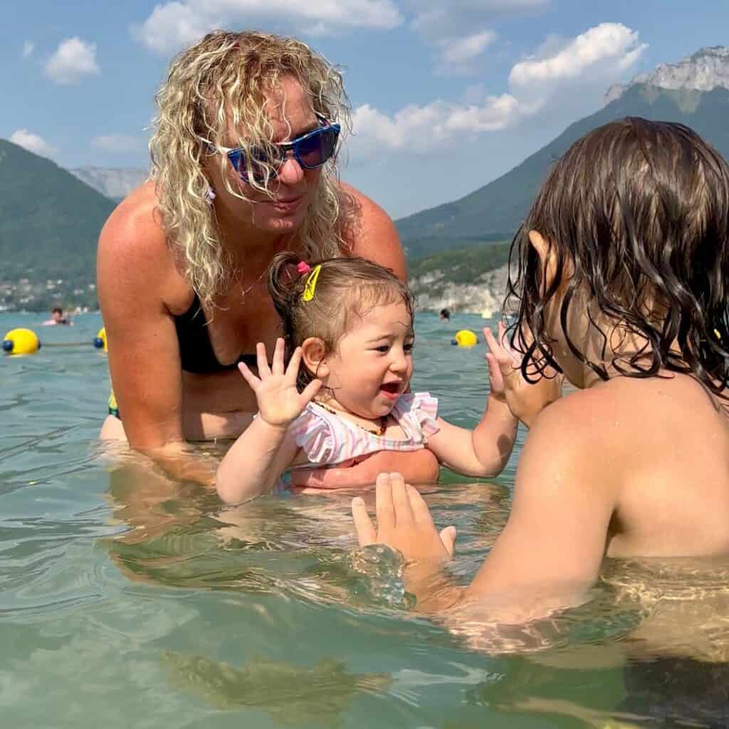 Woman and two children play together in the shallow water of Lake Annecy with mountains rising behind them in the French Alps. The lake is calm and clear on a warm summer afternoon in the Savoie region.