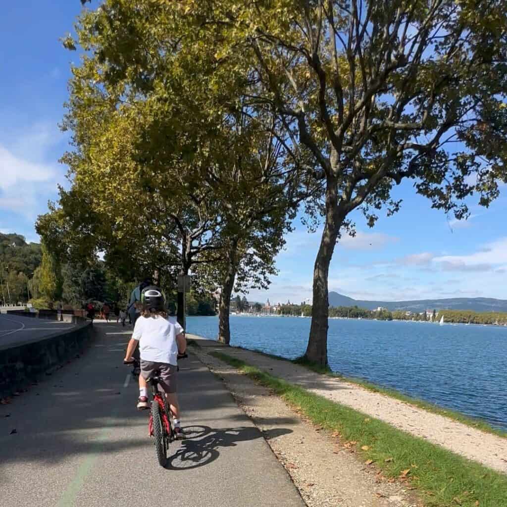 Child riding a bicycle along the lakeside cycling path beside Lake Annecy with tall trees casting shadows across the trail. Sailboats and mountains can be seen across the water on a sunny day in the French Alps.