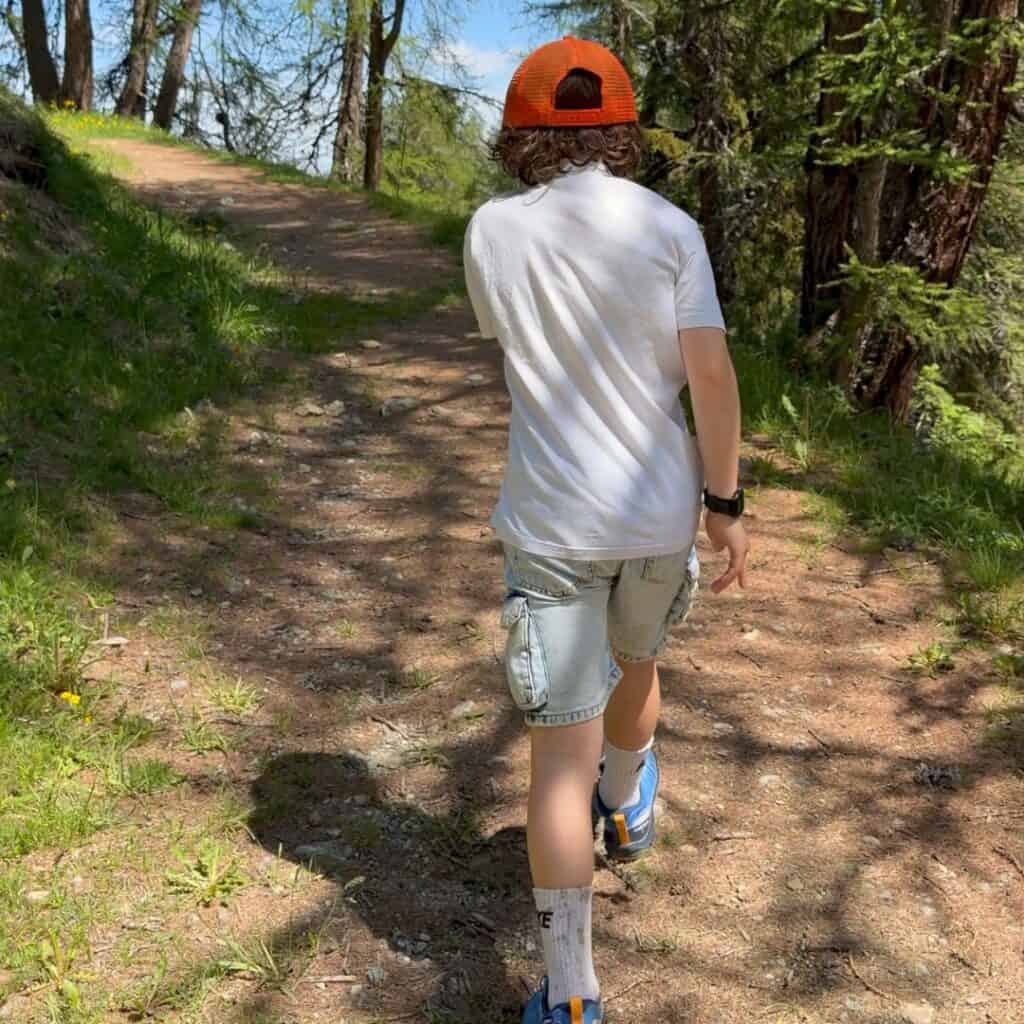 Child wearing an orange cap walks along a shaded forest trail surrounded by tall trees. The woodland path is suitable for a family-friendly summer walk in the mountains.