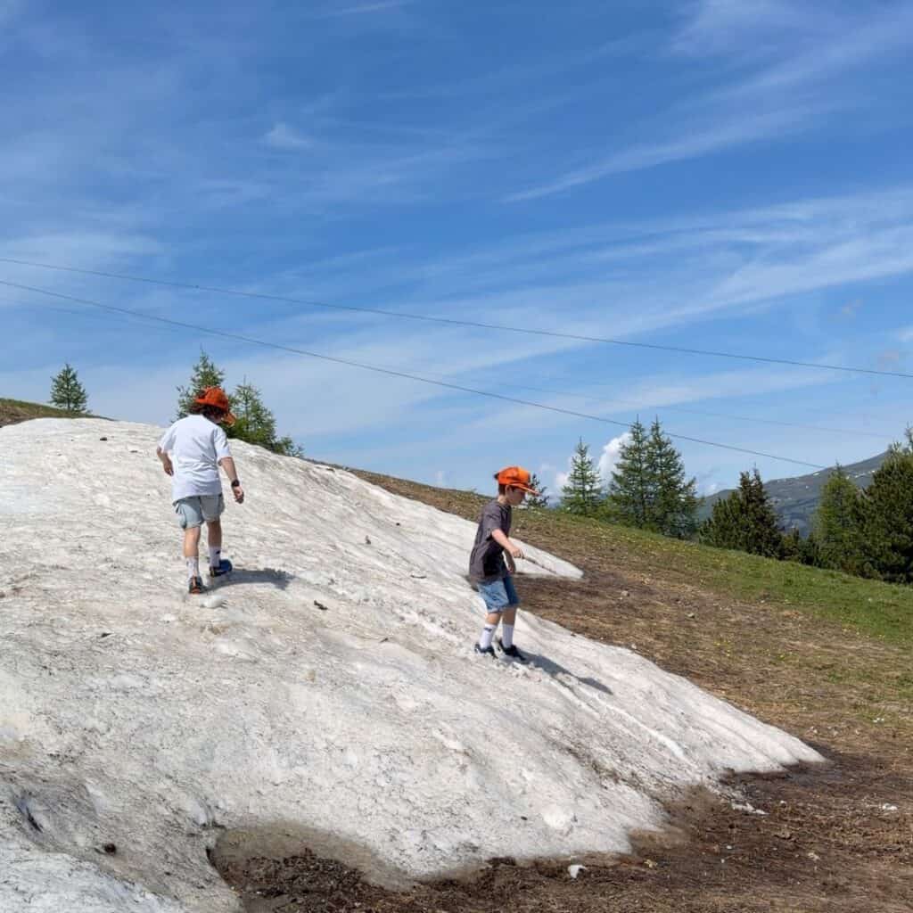 Two children in orange caps explore a lingering patch of snow on a mountain hillside during summer. The contrast between snow, green meadows and blue sky highlights the varied landscapes around La Plagne.