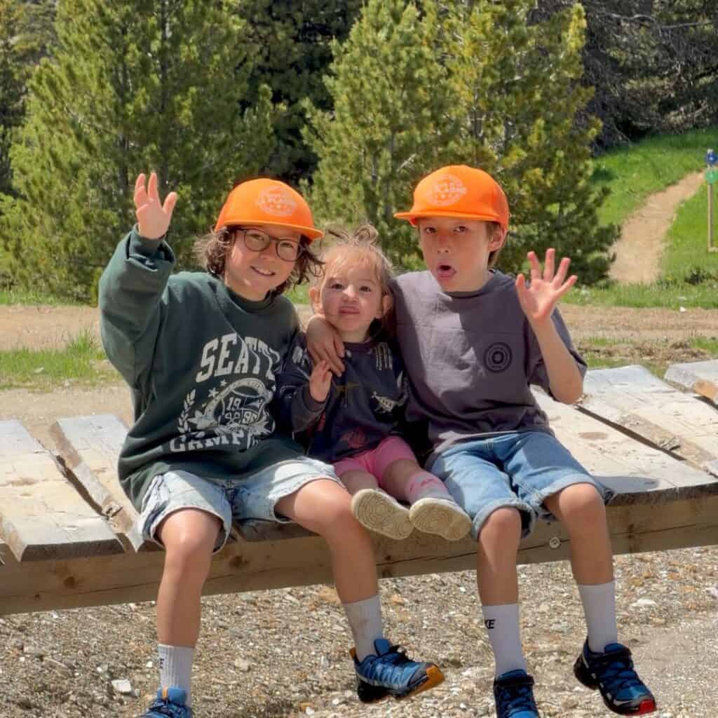 Three children wearing bright orange caps sit together on a wooden picnic table and wave at the camera. The rest area provides a fun stop during a family hiking day in the mountains.