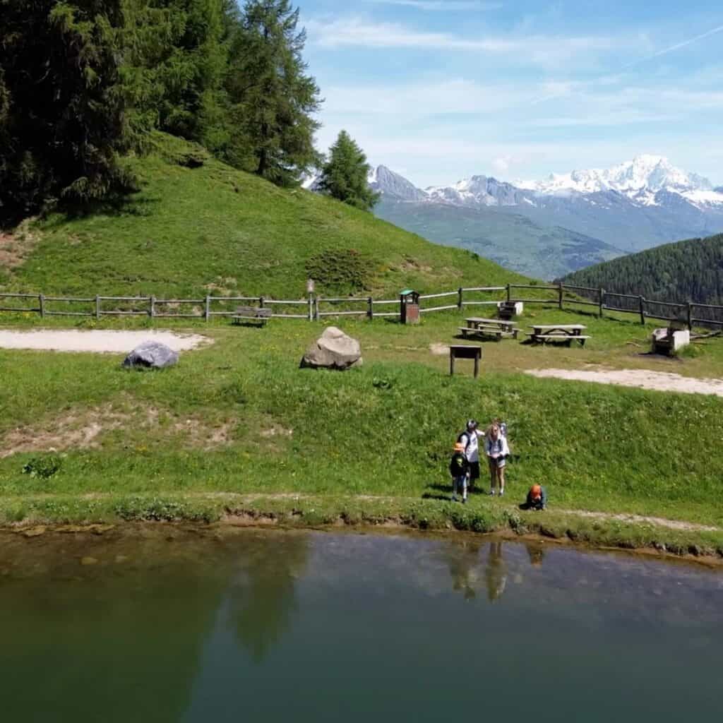 Family standing beside a calm mountain lake with snow-capped peaks rising in the distance. Picnic tables and open grassy slopes create a peaceful setting for a summer walk around La Plagne.