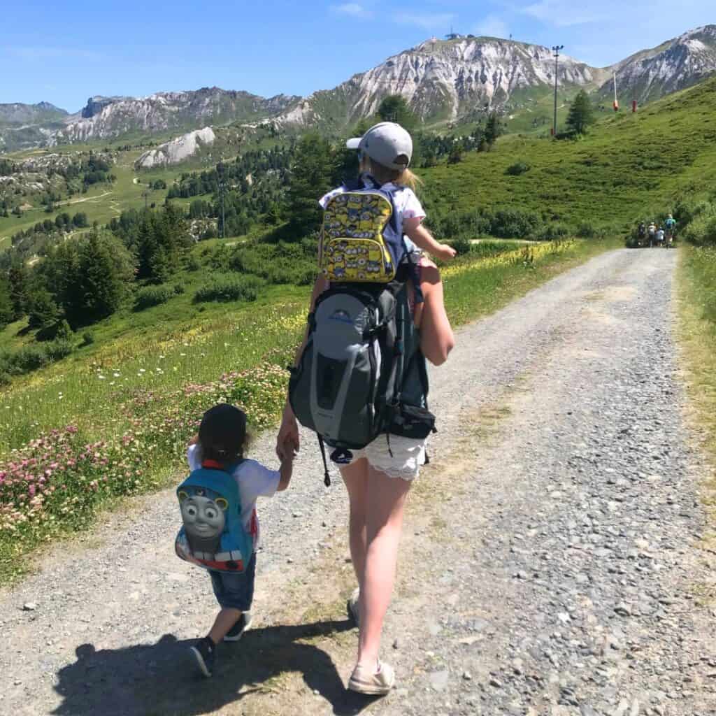 Adult carrying a toddler in a hiking backpack walks along a gravel mountain trail while holding a young child's hand. Wildflowers and dramatic alpine scenery line the route through La Plagne.