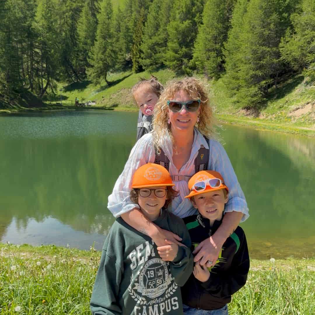 Family portrait beside a green mountain lake with forest reflected in the water. Two children in orange caps stand in front while a toddler rides in a backpack carrier, capturing a memorable day of family hiking.