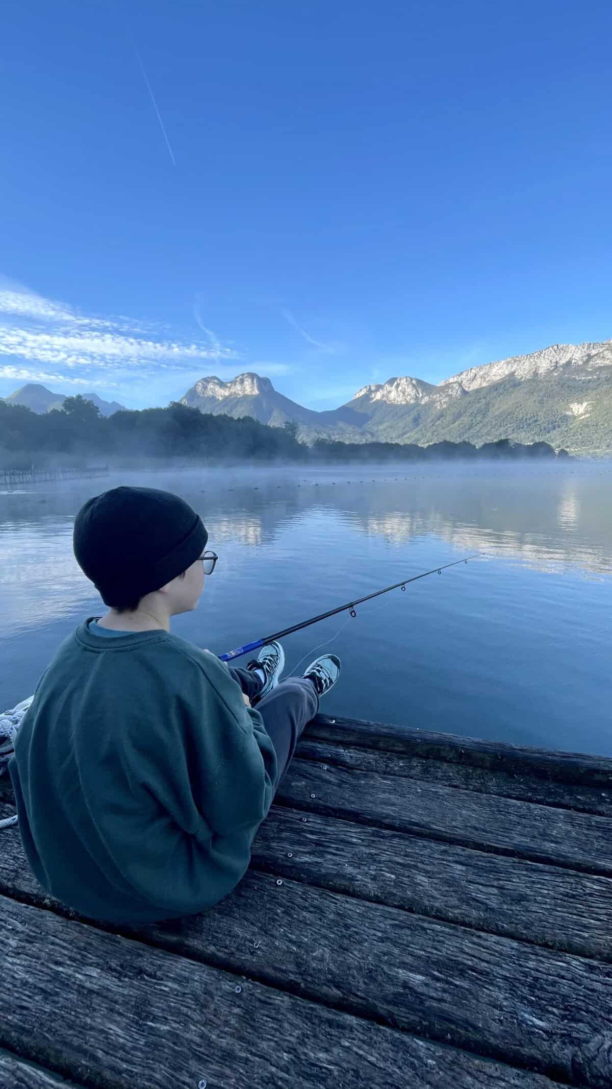 A young boy with a warm hat on say on a wooden jetty, fishing in the early morning haze at Lake Annecy