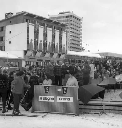 Black-and-white photo of crowds at the Interneige TV show in Plagne Centre, with a scoreboard reading La Plagne 9, Crans 2 and Bonnet Rouge banners on the building behind