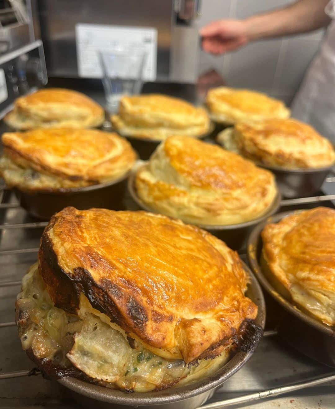 vegan restaurants la plagne Best vegan restaruants La Plagne: Freshly baked vegan pot pies with golden, flaky puff pastry tops, straight from the oven. The filling, visible where the pastry has expanded, appears creamy with vegetables and herbs. Multiple pies sit on a metal rack, with a blurred background showing a kitchen setting and a chef in a white uniform.